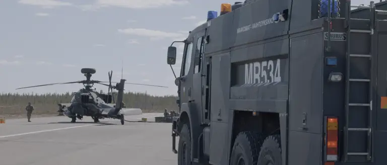 Image shows an AH64E Apache Helicopter stationary on the ground being refuelled by army air corps soldiers. A large Army vehicle is positioned on the right in the foreground.