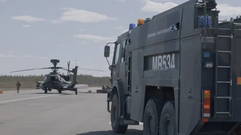 Image shows an AH64E Apache Helicopter stationary on the ground being refuelled by army air corps soldiers. A large Army vehicle is positioned on the right in the foreground.
