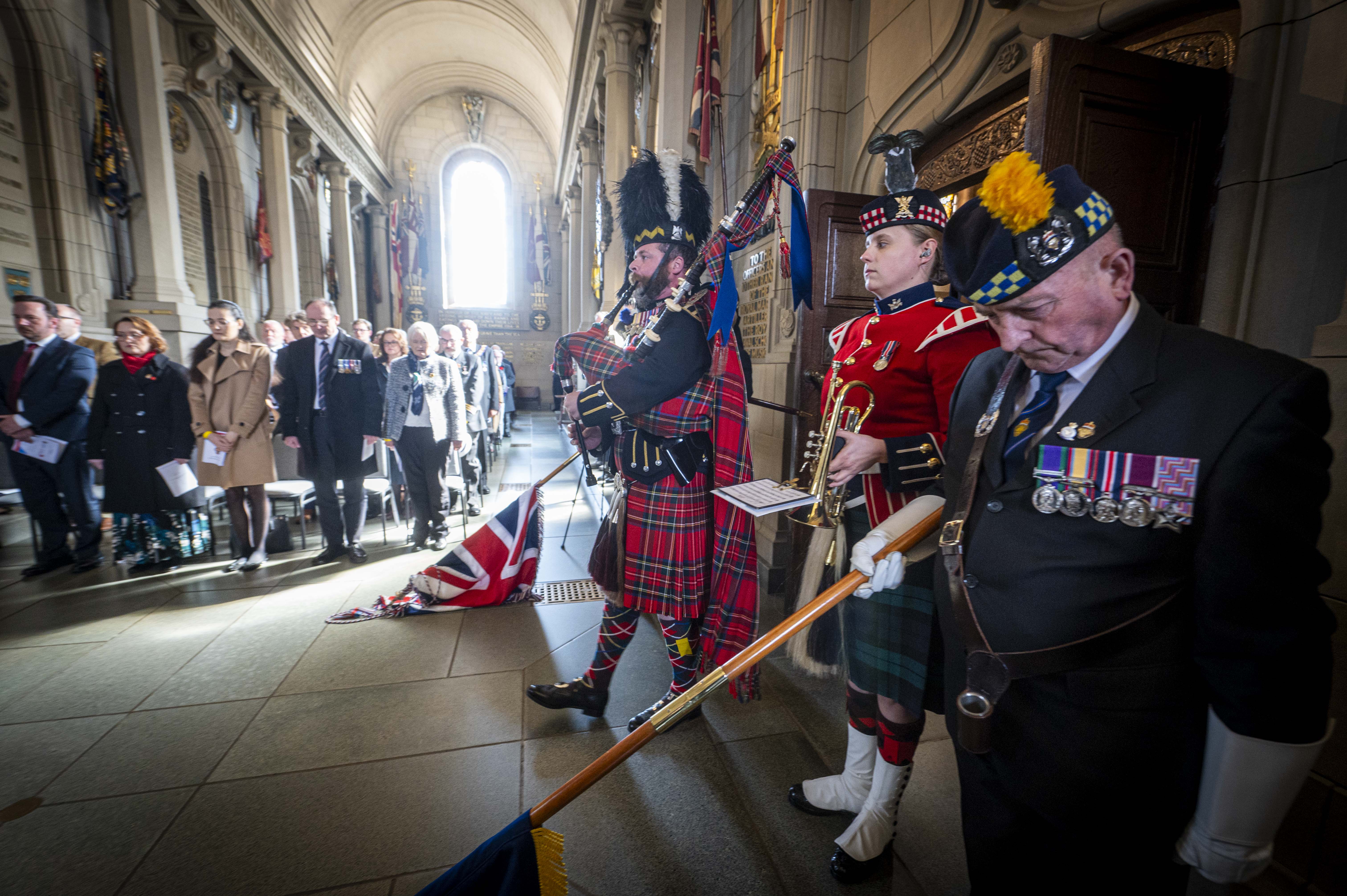 A bagpiper plays the pipes. He is wearing a tall black bearskin hat, and red tartan. Either side of him, people lower flags.