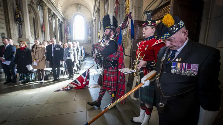 A bagpiper plays the pipes. He is wearing a tall black bearskin hat, and red tartan. Either side of him, people lower flags.