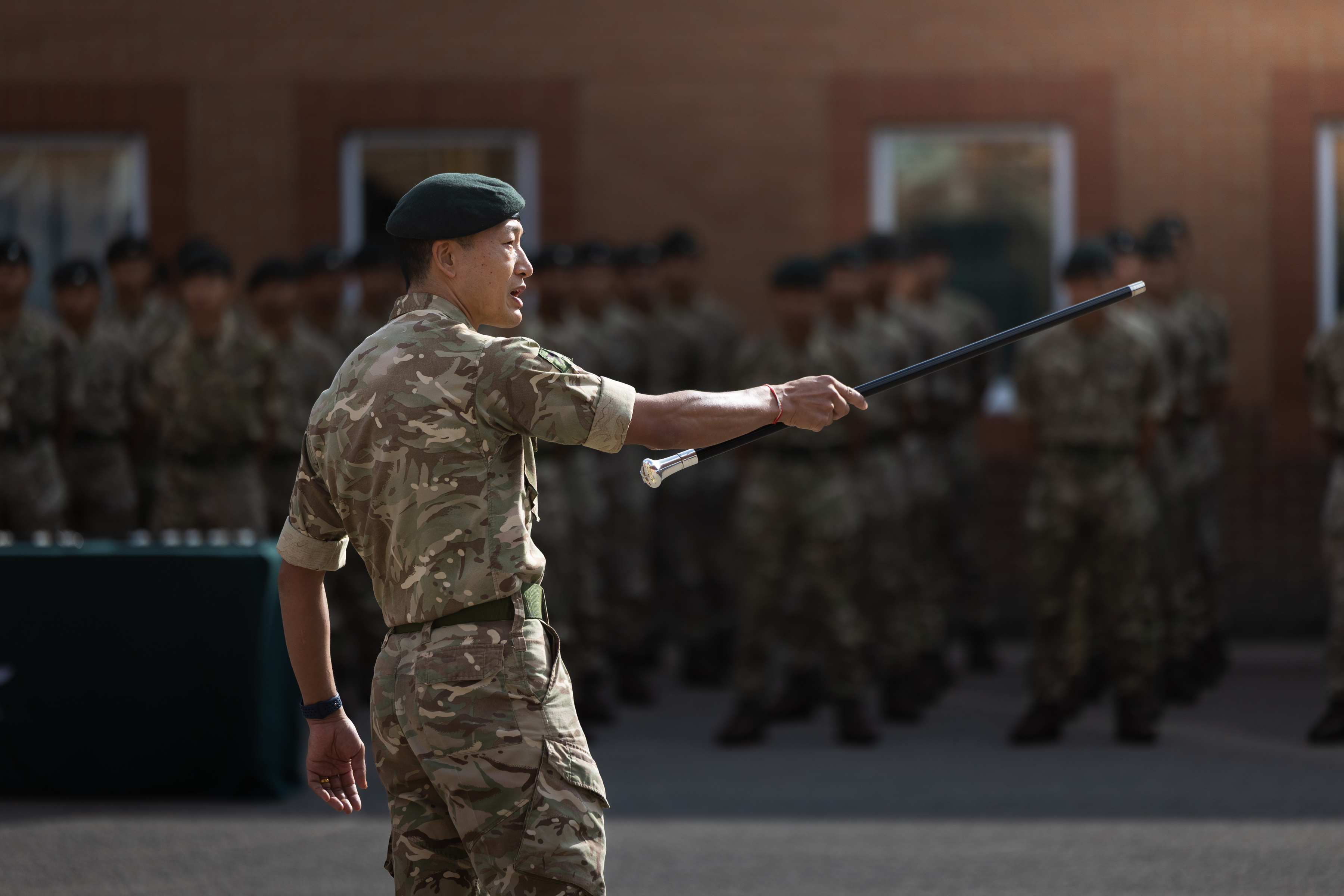 Gurkha instructor with long black stick pointed towards rows of soldiers outside on a parade square. Everybody is wearing camouflage uniform.