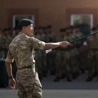 Gurkha instructor with long black stick pointed towards rows of soldiers outside on a parade square. Everybody is wearing camouflage uniform.