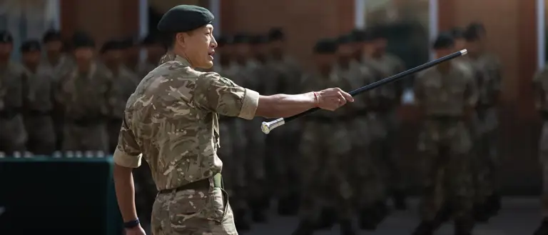 Gurkha instructor with long black stick pointed towards rows of soldiers outside on a parade square. Everybody is wearing camouflage uniform.