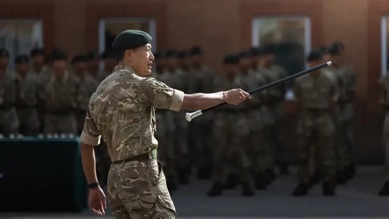 Gurkha instructor with long black stick pointed towards rows of soldiers outside on a parade square. Everybody is wearing camouflage uniform.