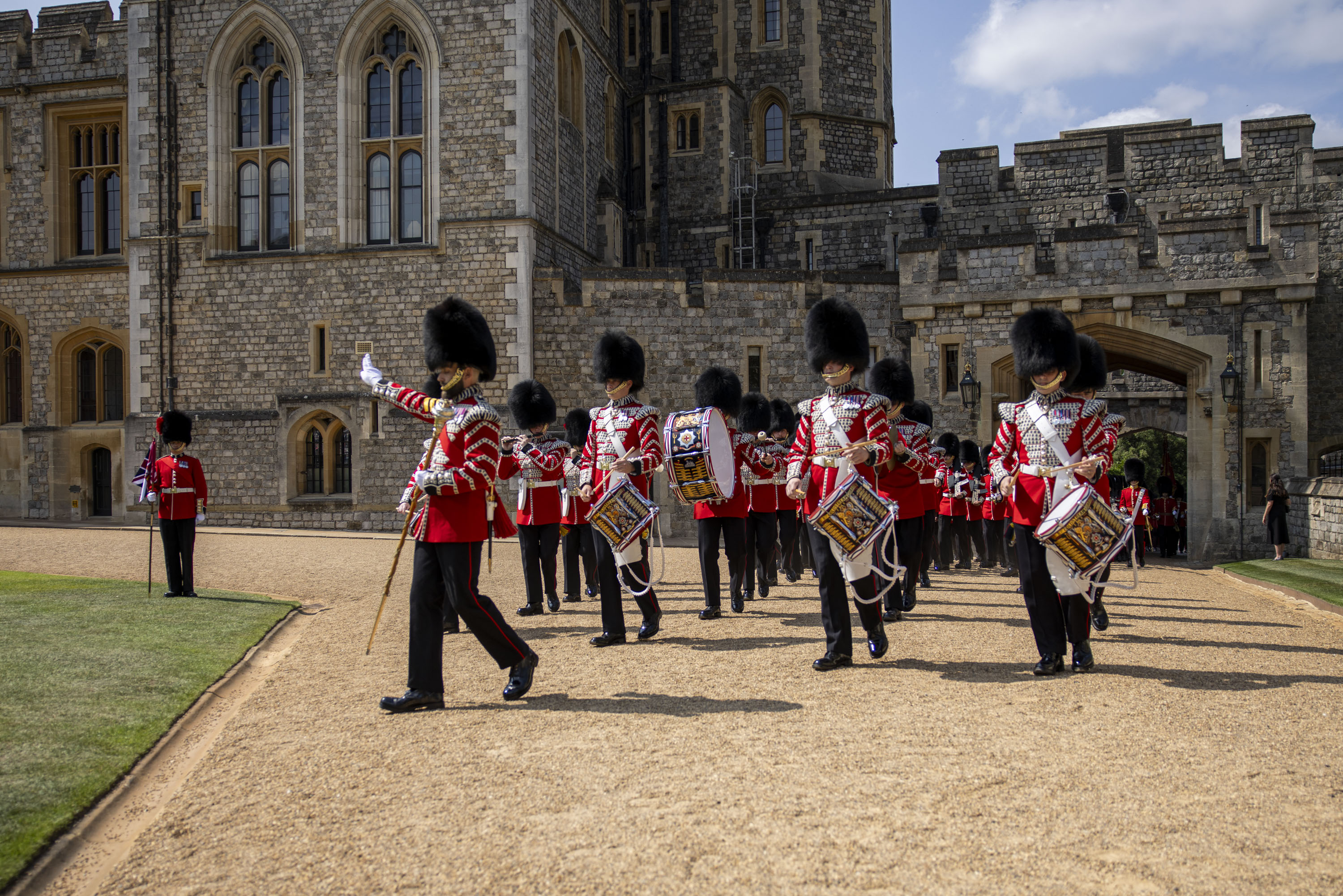 Soldiers wearing red tunics and bearskin hats march whilst playing the drums. 
