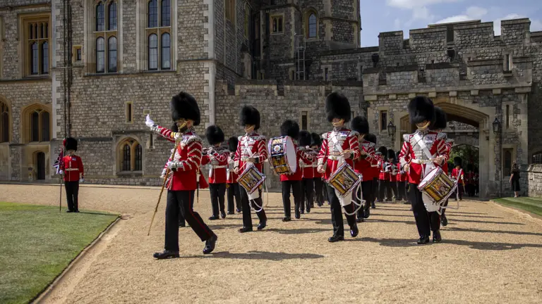 Soldiers wearing red tunics and bearskin hats march whilst playing the drums.