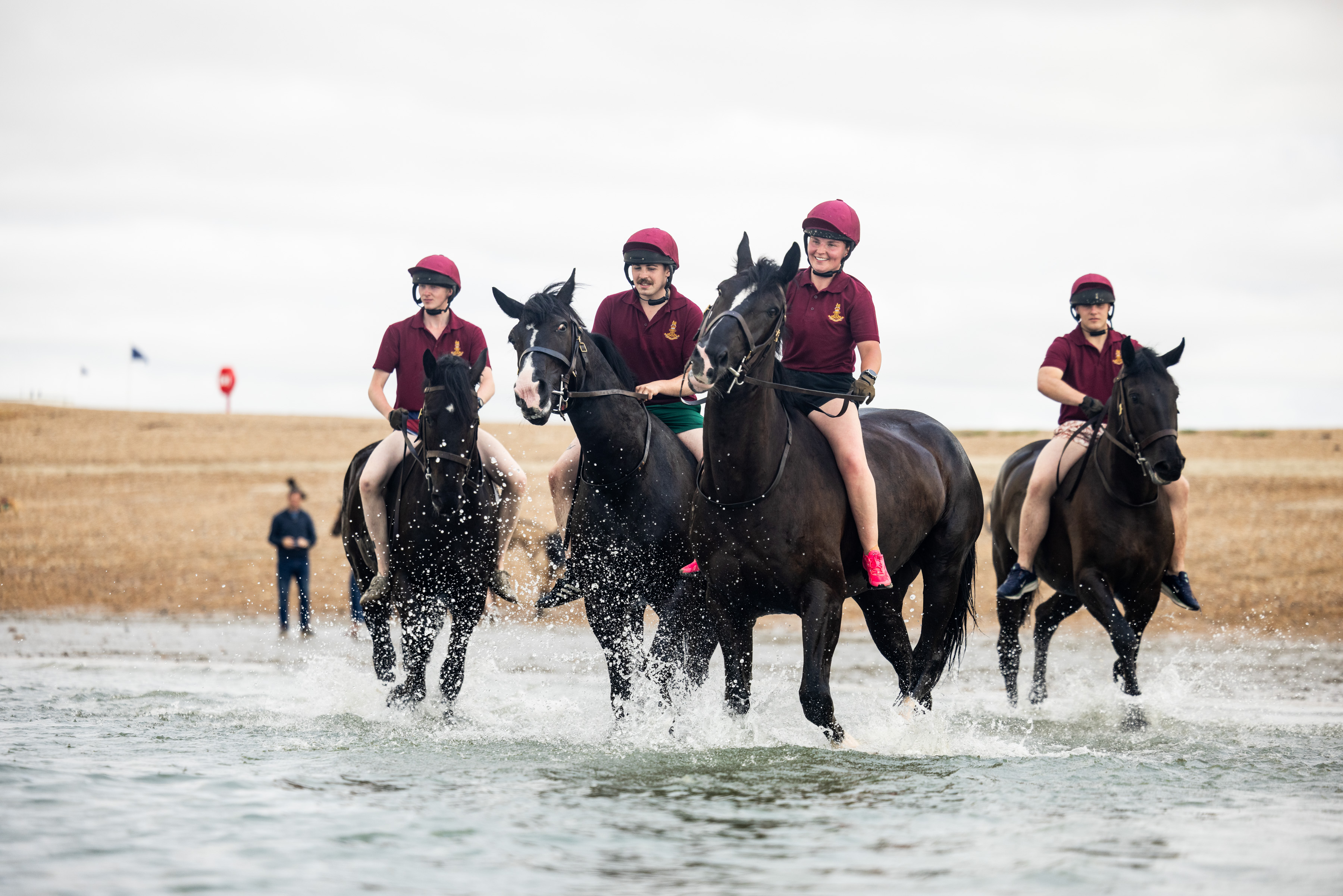 A group of individuals riding dark brown horses through shallow ocean water. They wear maroon shirts and helmets, with one rider holding a baton. The shoreline is visible, and a person stands in the distance.