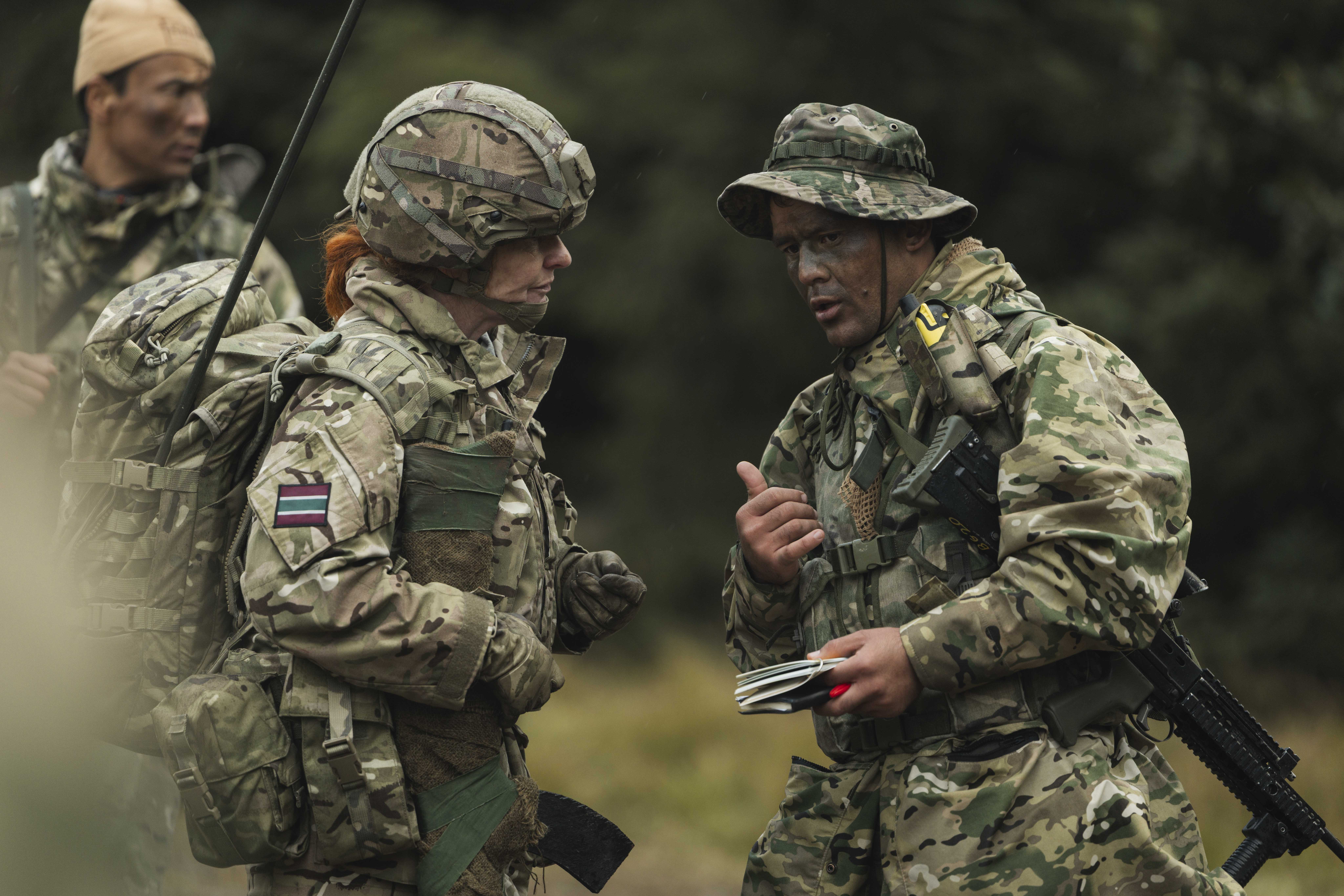 Three soldiers in camouflage uniforms are engaged in discussion in an outdoor setting. Two are standing close together.
