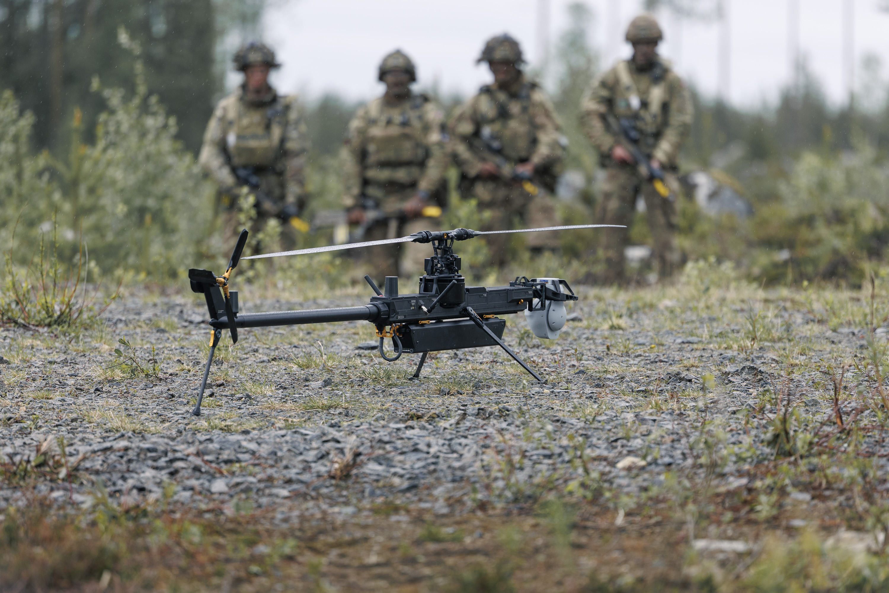 Two individuals in military gear are outdoors in a forested area, focusing on a small unmanned aircraft system on the ground. One is kneeling and adjusting the drone, while the other stands nearby holding equipment. The scene is set on a gravel surface with trees in the background.