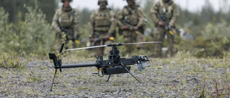 Two individuals in military gear are outdoors in a forested area, focusing on a small unmanned aircraft system on the ground. One is kneeling and adjusting the drone, while the other stands nearby holding equipment. The scene is set on a gravel surface with trees in the background.