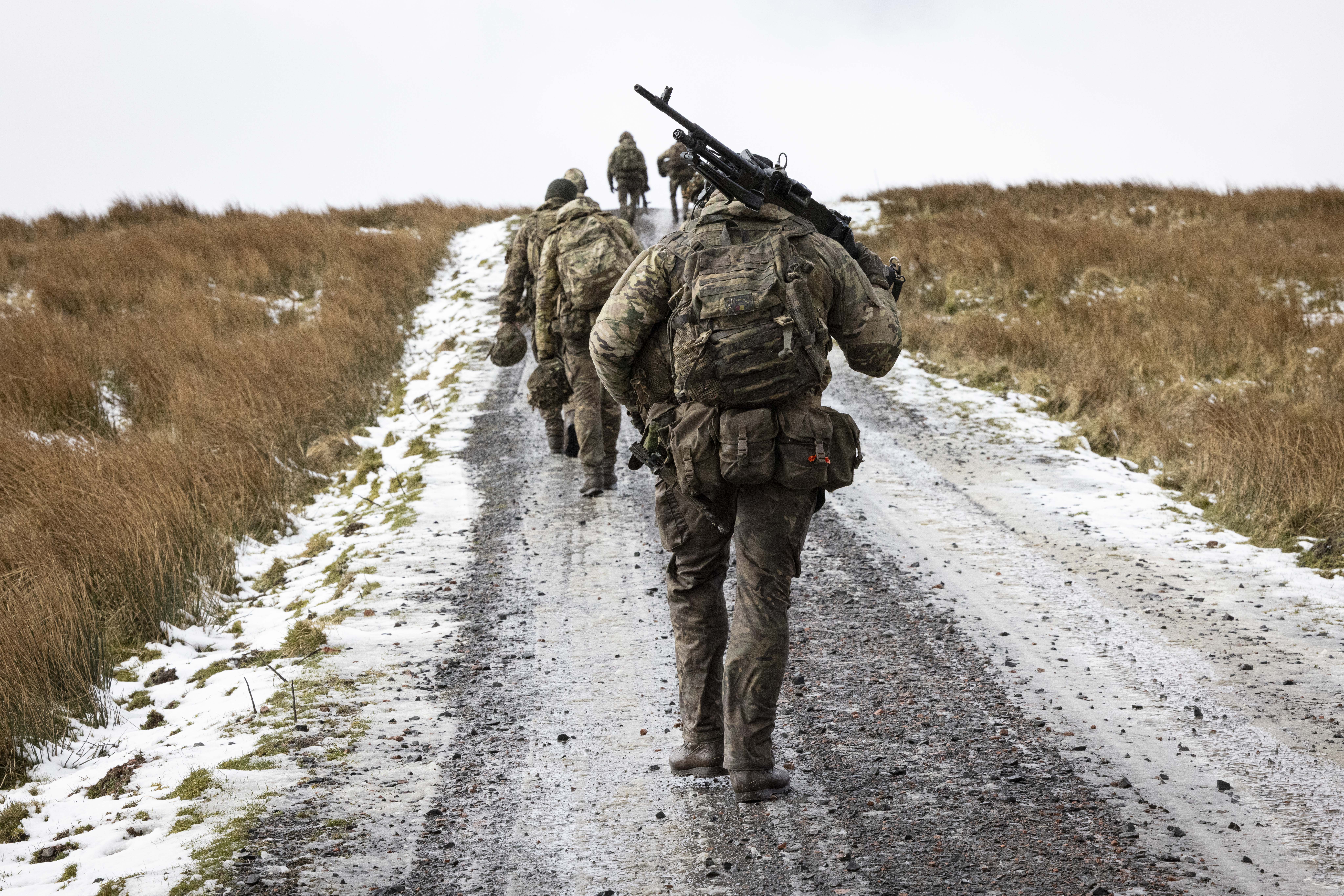 Soldiers in camouflage uniform wearing body armour and helmet walks in the firing range carrying their equipment as they walk up hill with snow on the ground. 
