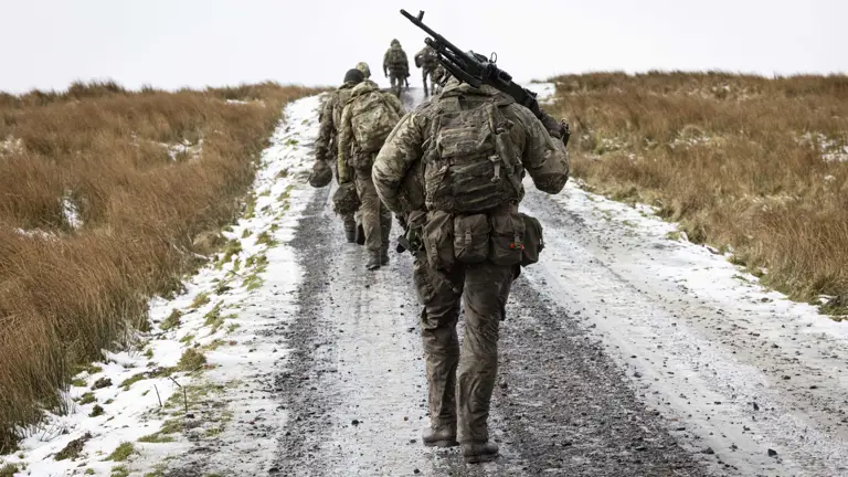 Soldiers in camouflage uniform wearing body armour and helmet walks in the firing range carrying their equipment as they walk up hill with snow on the ground.