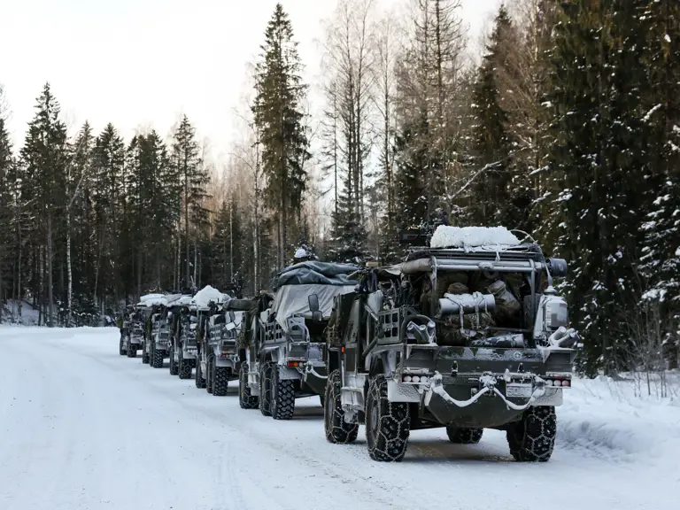 A convoy of military vehicles covered in snow parked along a snowy forest road during winter.