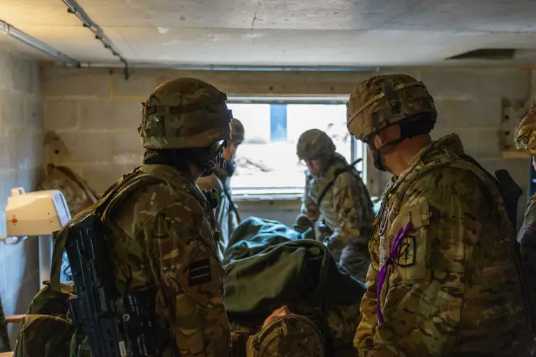 Soldiers in camouflage gear attending to a covered stretcher inside a concrete room with a small window.