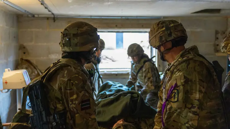 Soldiers in camouflage gear attending to a covered stretcher inside a concrete room with a small window.