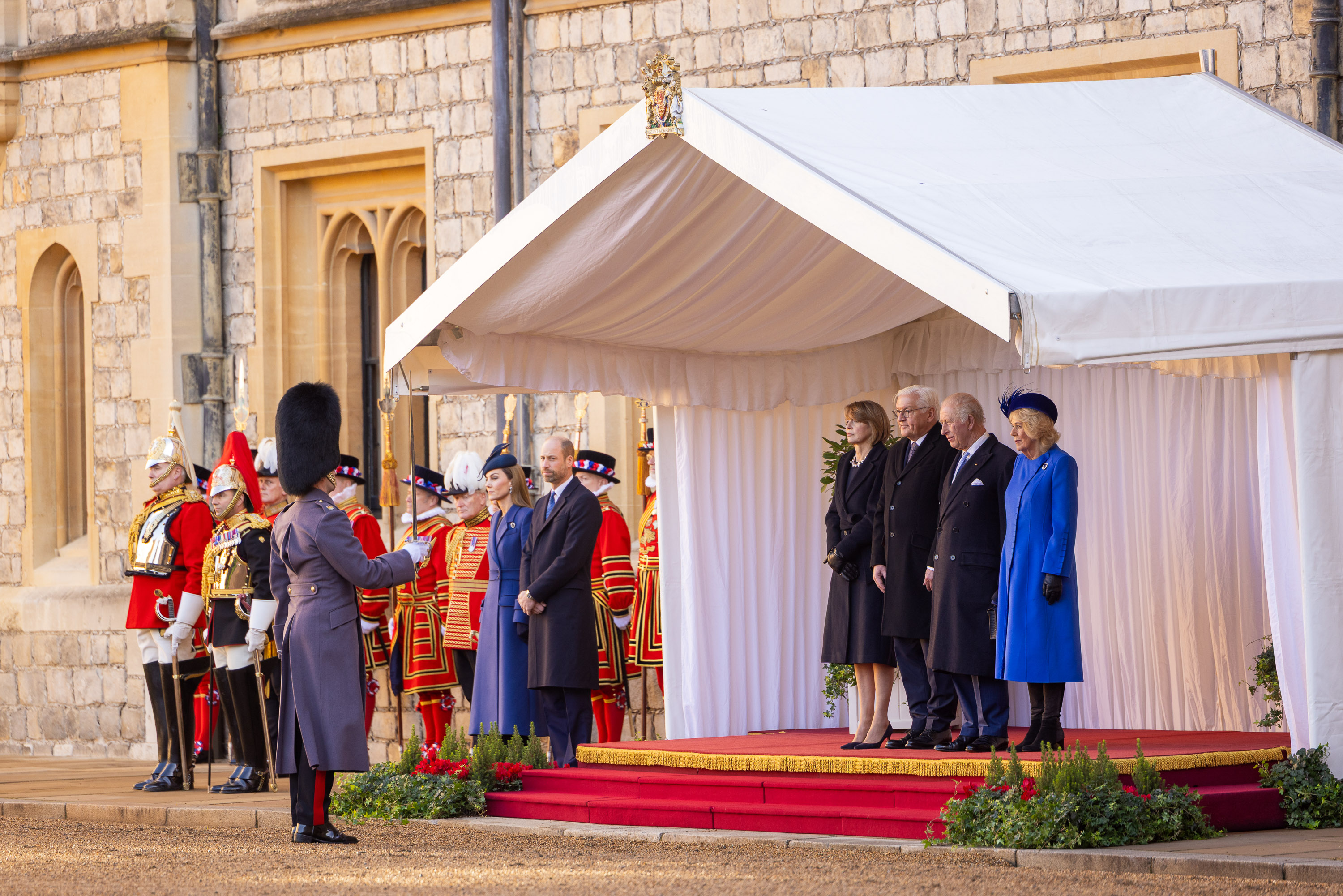 A formal ceremony is taking place outdoors. People stand on a red-carpeted platform under a white canopy, with guards dressed in ceremonial uniforms nearby.