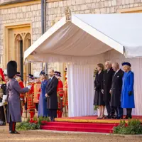 A formal ceremony is taking place outdoors. People stand on a red-carpeted platform under a white canopy, with guards dressed in ceremonial uniforms nearby.