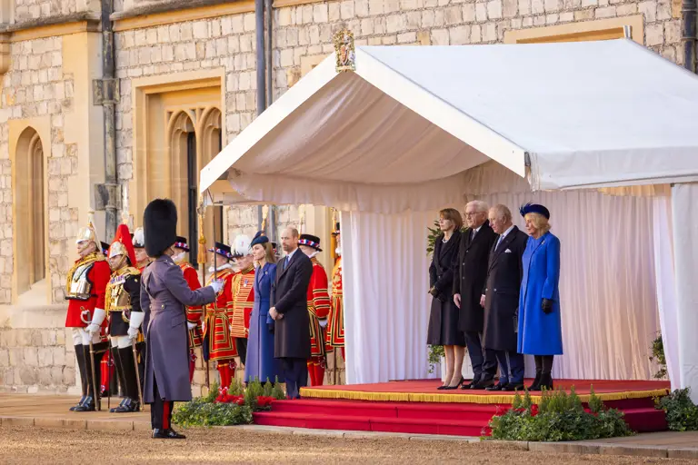 A formal ceremony is taking place outdoors. People stand on a red-carpeted platform under a white canopy, with guards dressed in ceremonial uniforms nearby.