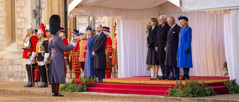 A formal ceremony is taking place outdoors. People stand on a red-carpeted platform under a white canopy, with guards dressed in ceremonial uniforms nearby.