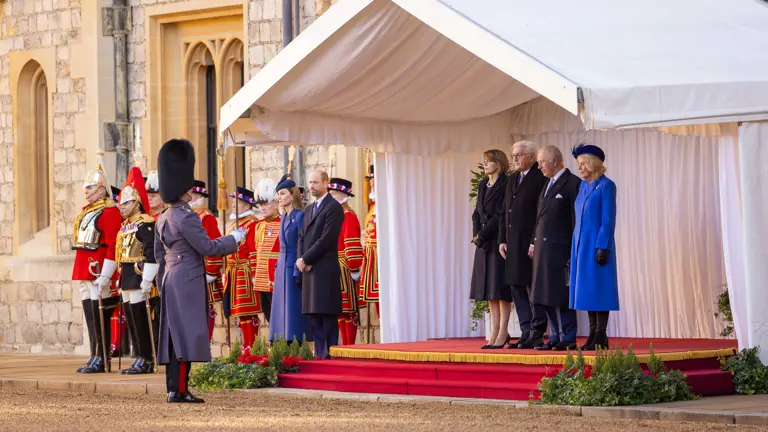 A formal ceremony is taking place outdoors. People stand on a red-carpeted platform under a white canopy, with guards dressed in ceremonial uniforms nearby.