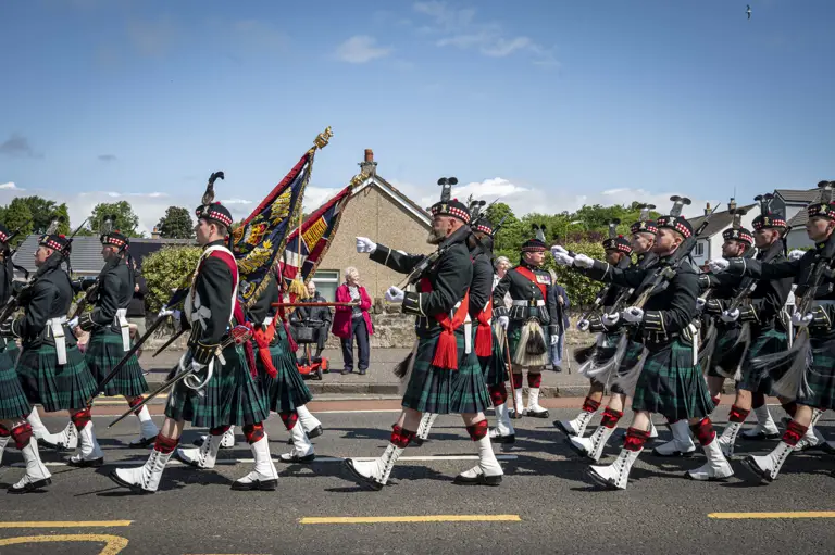 Men wearing black tunics and green and blue kilts march past. They are carrying rifles.