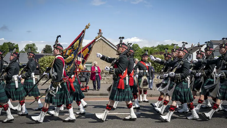 Men wearing black tunics and green and blue kilts march past. They are carrying rifles.