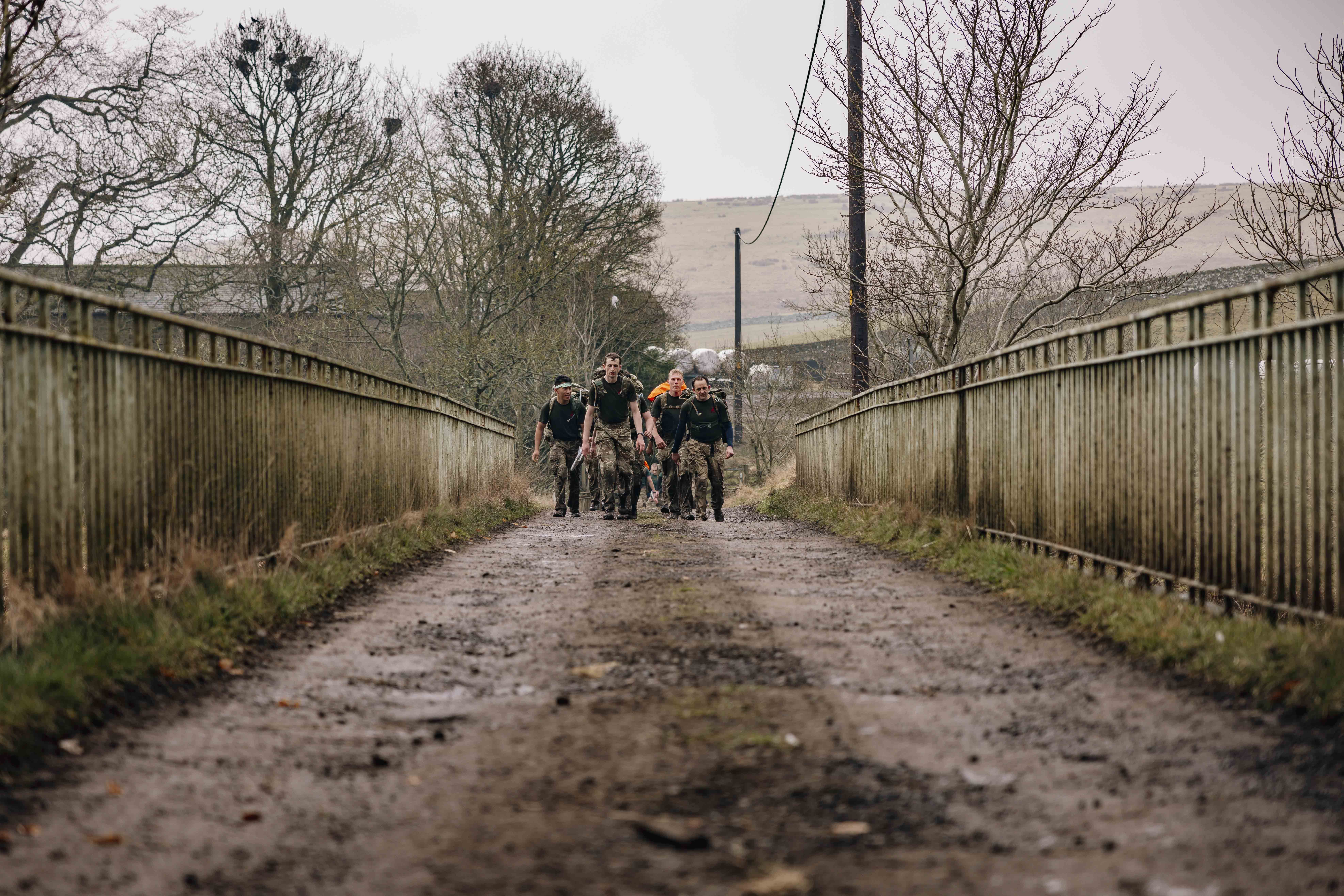 Soldier are seen carrying their gear in large green camouflage backpacks walking over a bridge in the Otterburn countryside in the wet weather. 