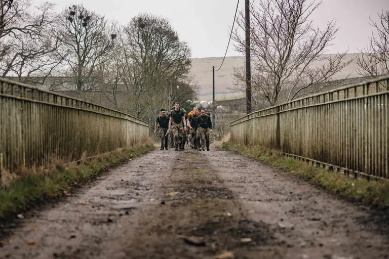 Soldier are seen carrying their gear in large green camouflage backpacks walking over a bridge in the Otterburn countryside in the wet weather.