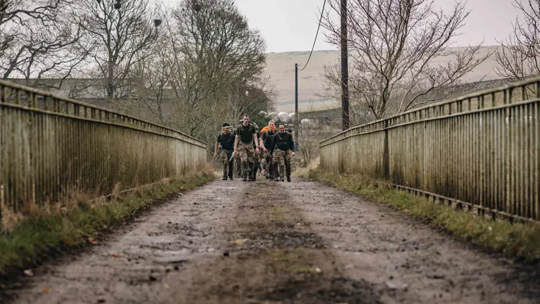 Soldier are seen carrying their gear in large green camouflage backpacks walking over a bridge in the Otterburn countryside in the wet weather.