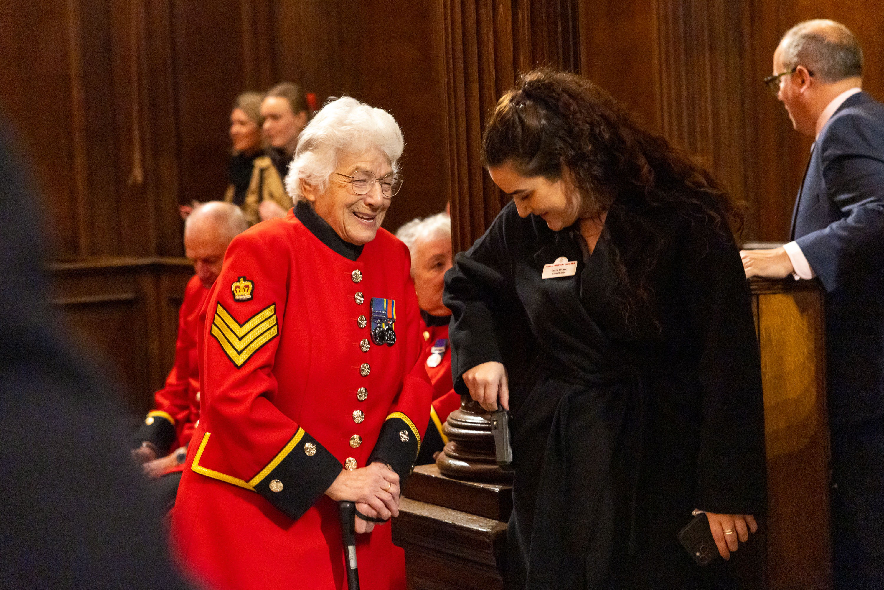 Elderly woman in a red ceremonial uniform with medals talks to a woman in black inside a wood-panelled room.