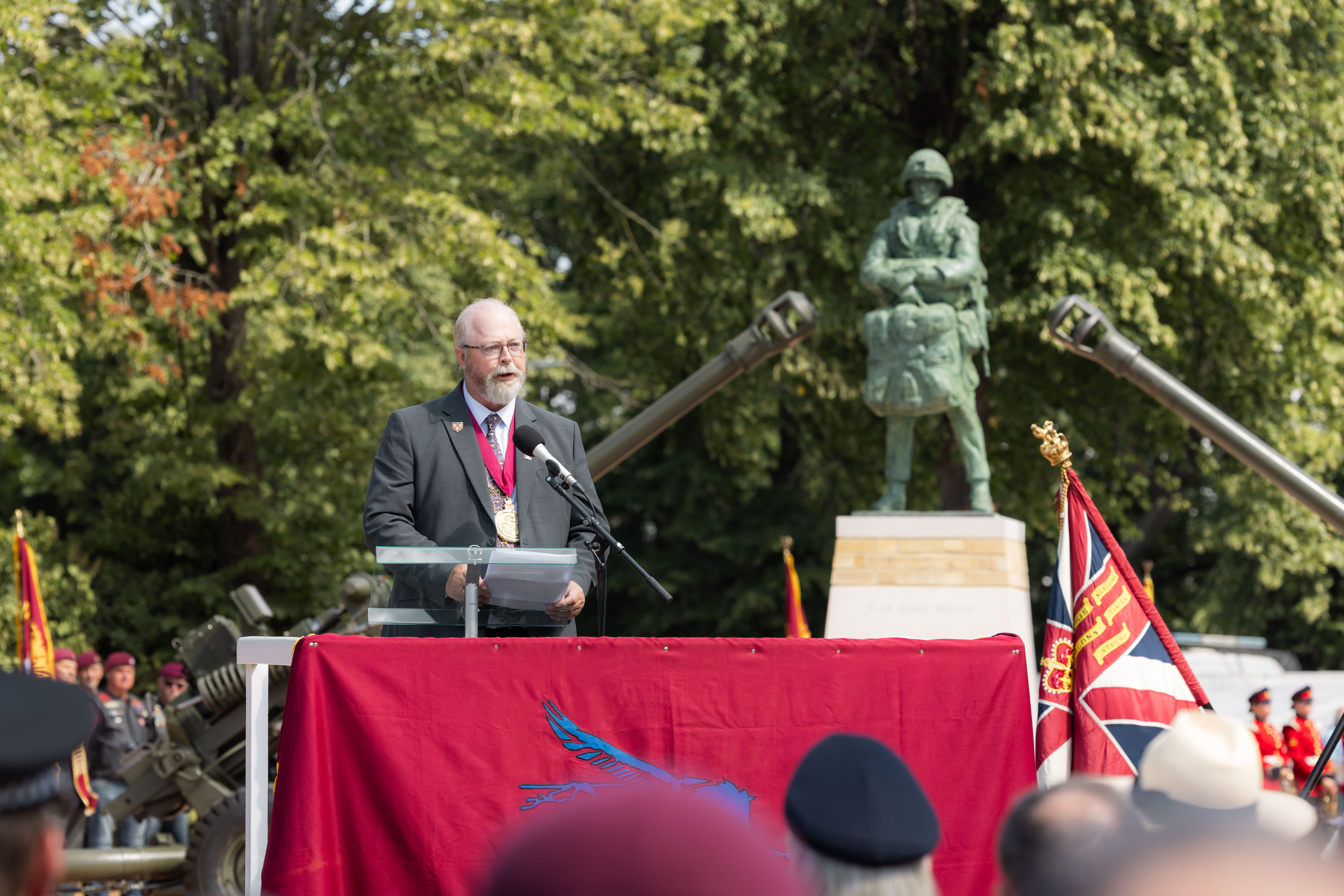 Man in suit stood behind a red banner talking to a crowd, the bronze statue behind him between two big guns.