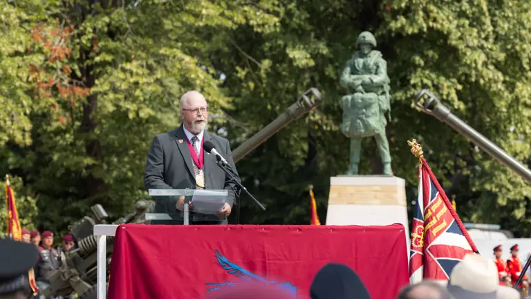 Man in suit stood behind a red banner talking to a crowd, the bronze statue behind him between two big guns.