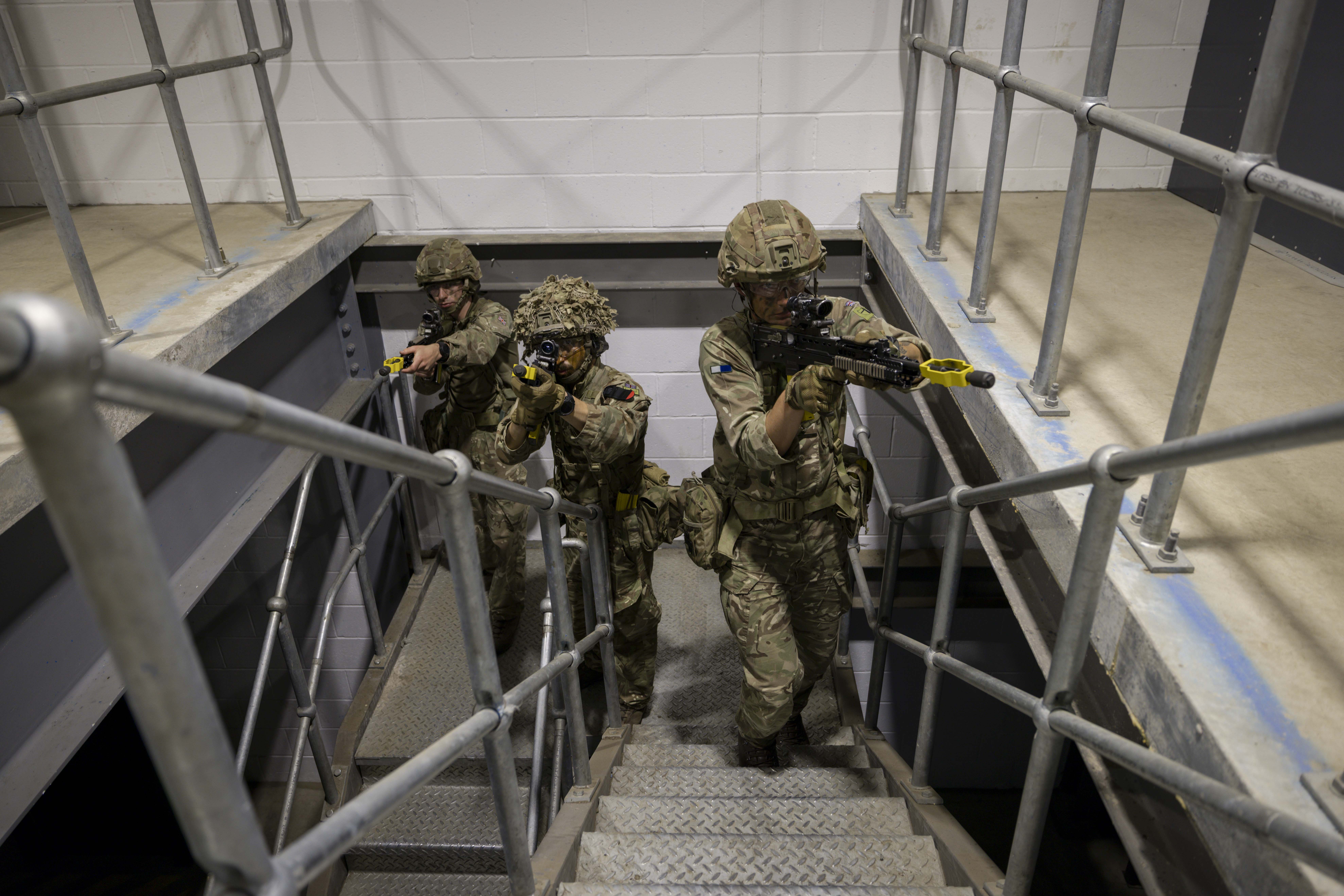 Soldiers in camouflage uniforms holding rifles stand ready in concrete building on metal stairs.