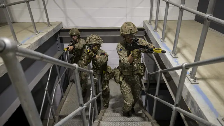 Soldiers in camouflage uniforms holding rifles stand ready in concrete building on metal stairs.