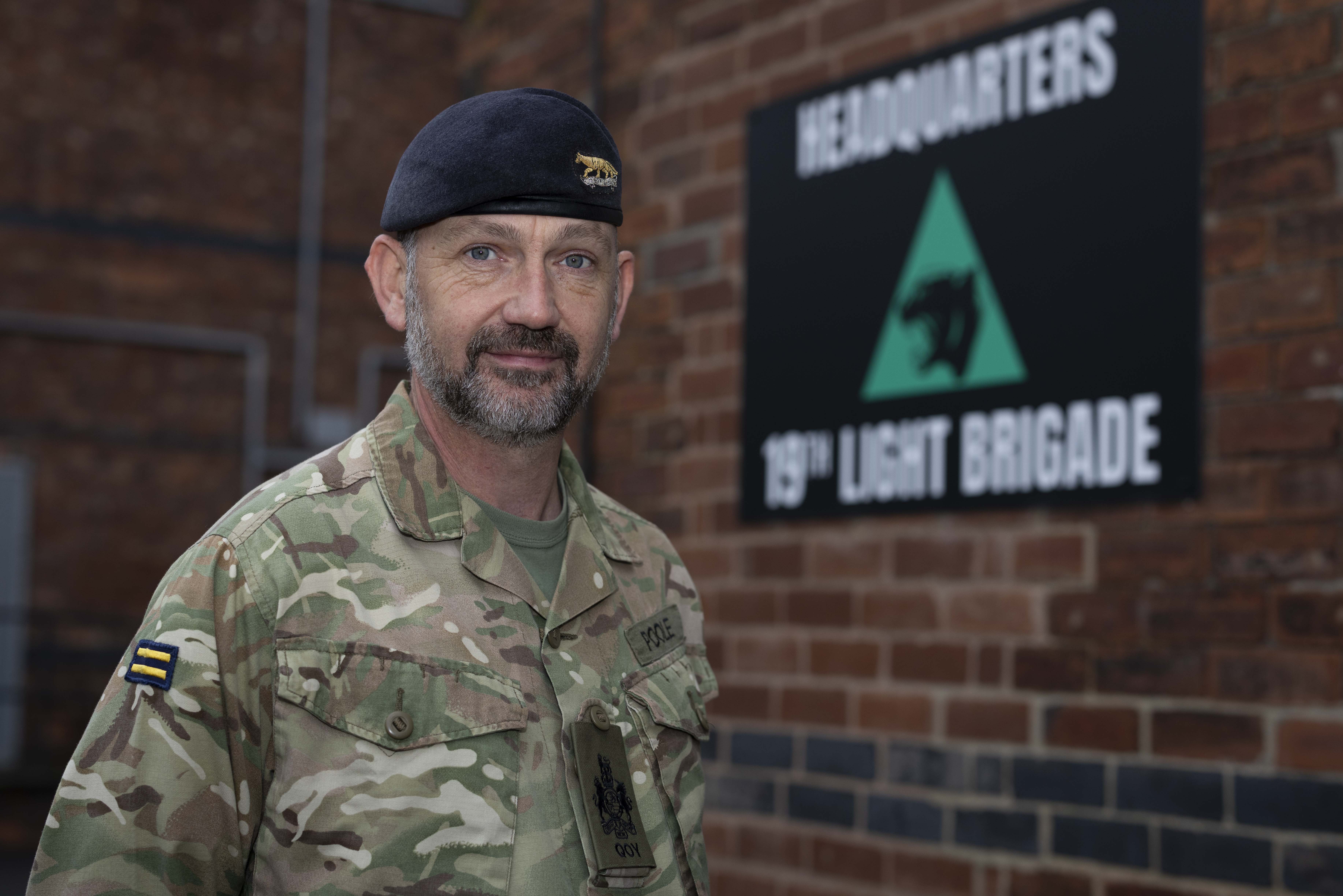 Soldier in camouflage uniform standing in front of a brick wall with a 19th Light Brigade headquarters sign.