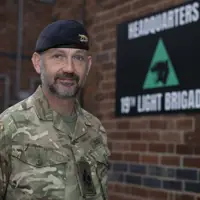 Soldier in camouflage uniform standing in front of a brick wall with a 19th Light Brigade headquarters sign.