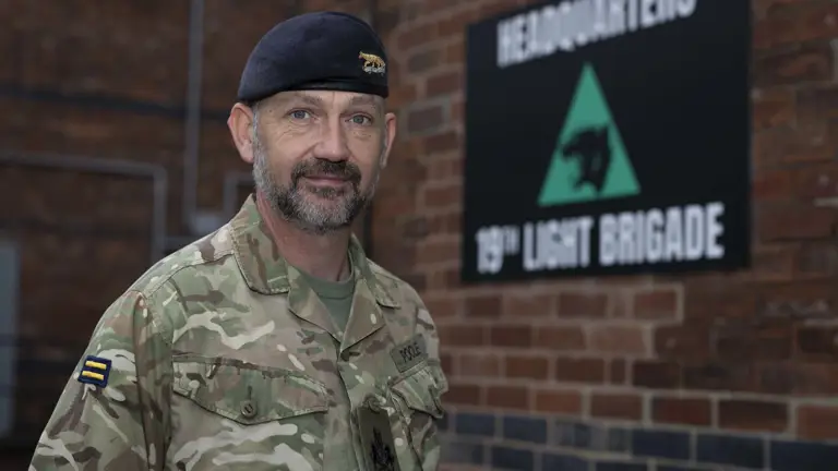 Soldier in camouflage uniform standing in front of a brick wall with a 19th Light Brigade headquarters sign.