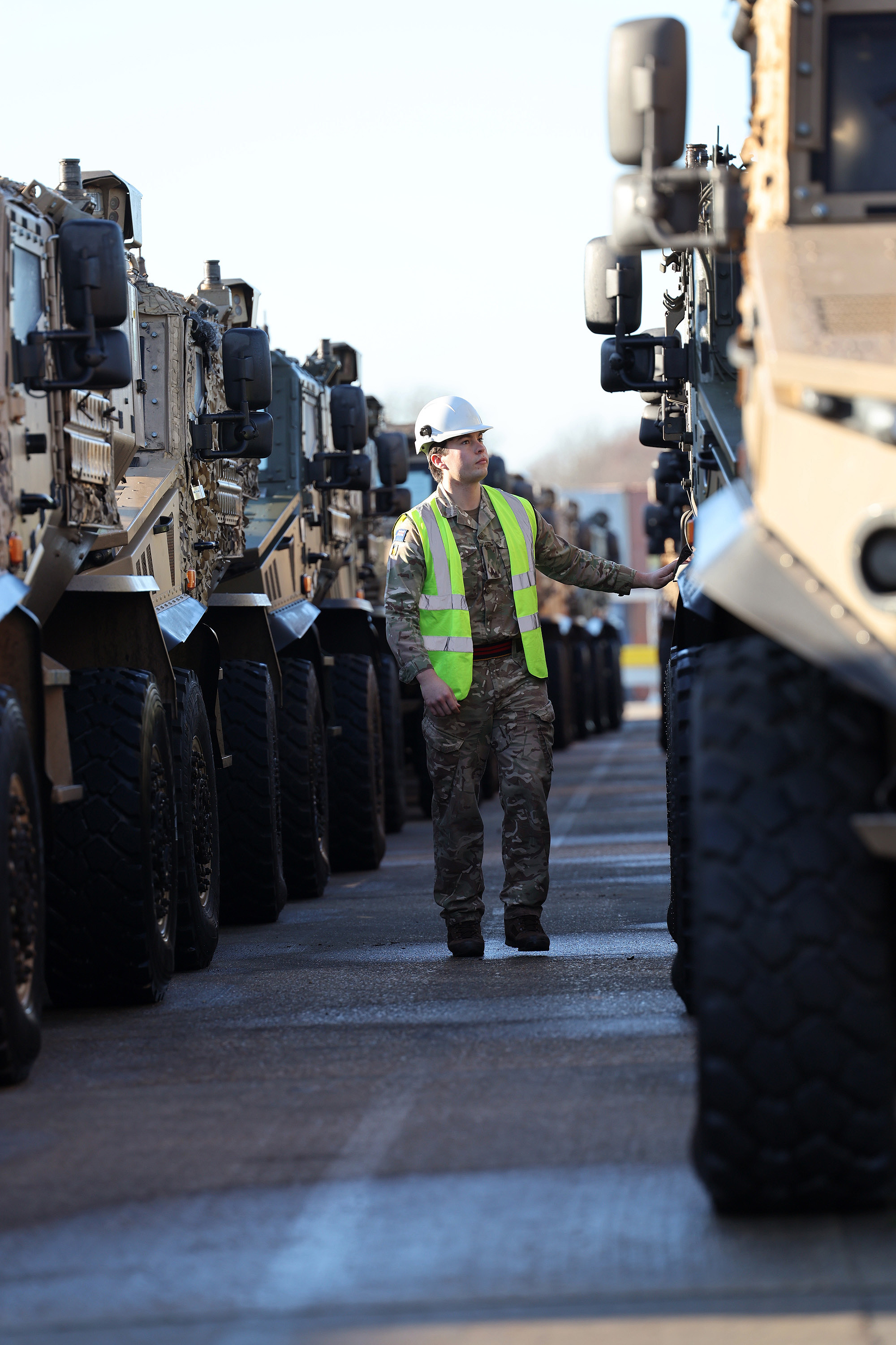 Soldier in camouflage uniform is seen walking between lined up vehicles while wearing a hi vis jacket and a hard hat checking vehicles over before transportation.