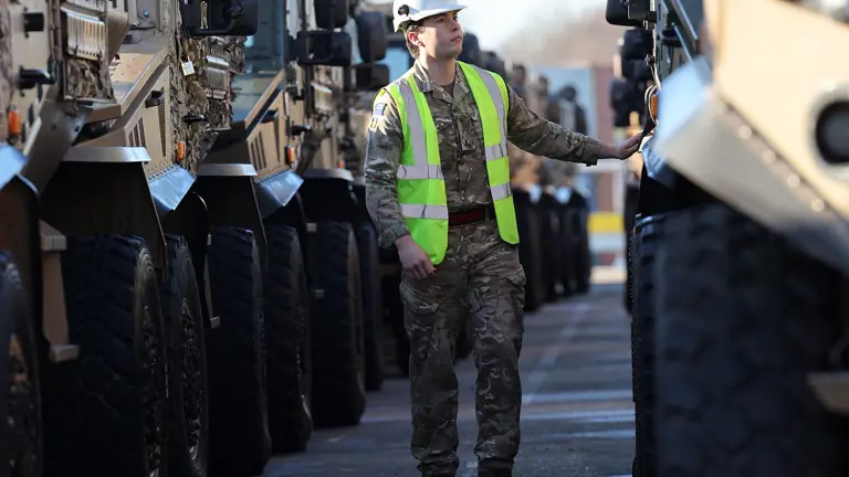 Soldier in camouflage uniform is seen walking between lined up vehicles while wearing a hi vis jacket and a hard hat checking vehicles over before transportation.