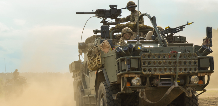 A green armoured vehicle drives across sand.