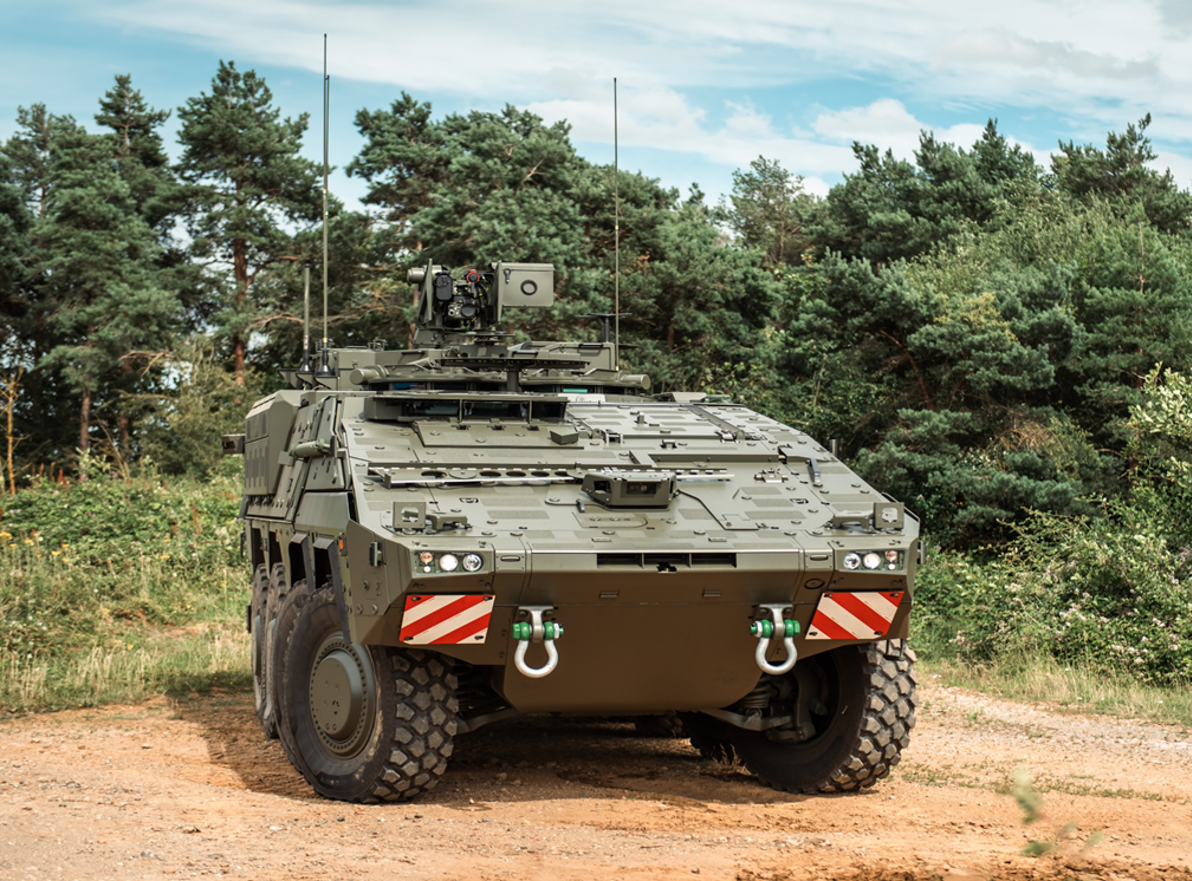 A military armoured vehicle is parked on a dirt path surrounded by trees, under a partly cloudy sky. 