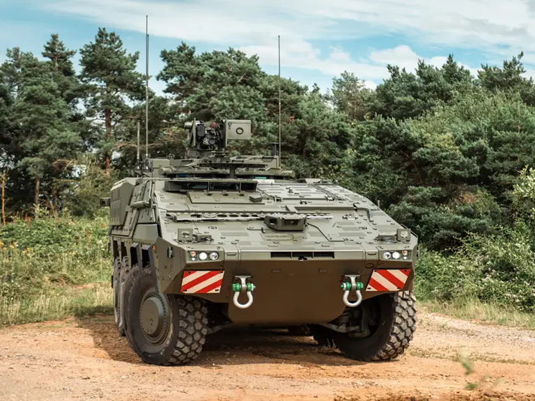 A military armoured vehicle is parked on a dirt path surrounded by trees, under a partly cloudy sky.