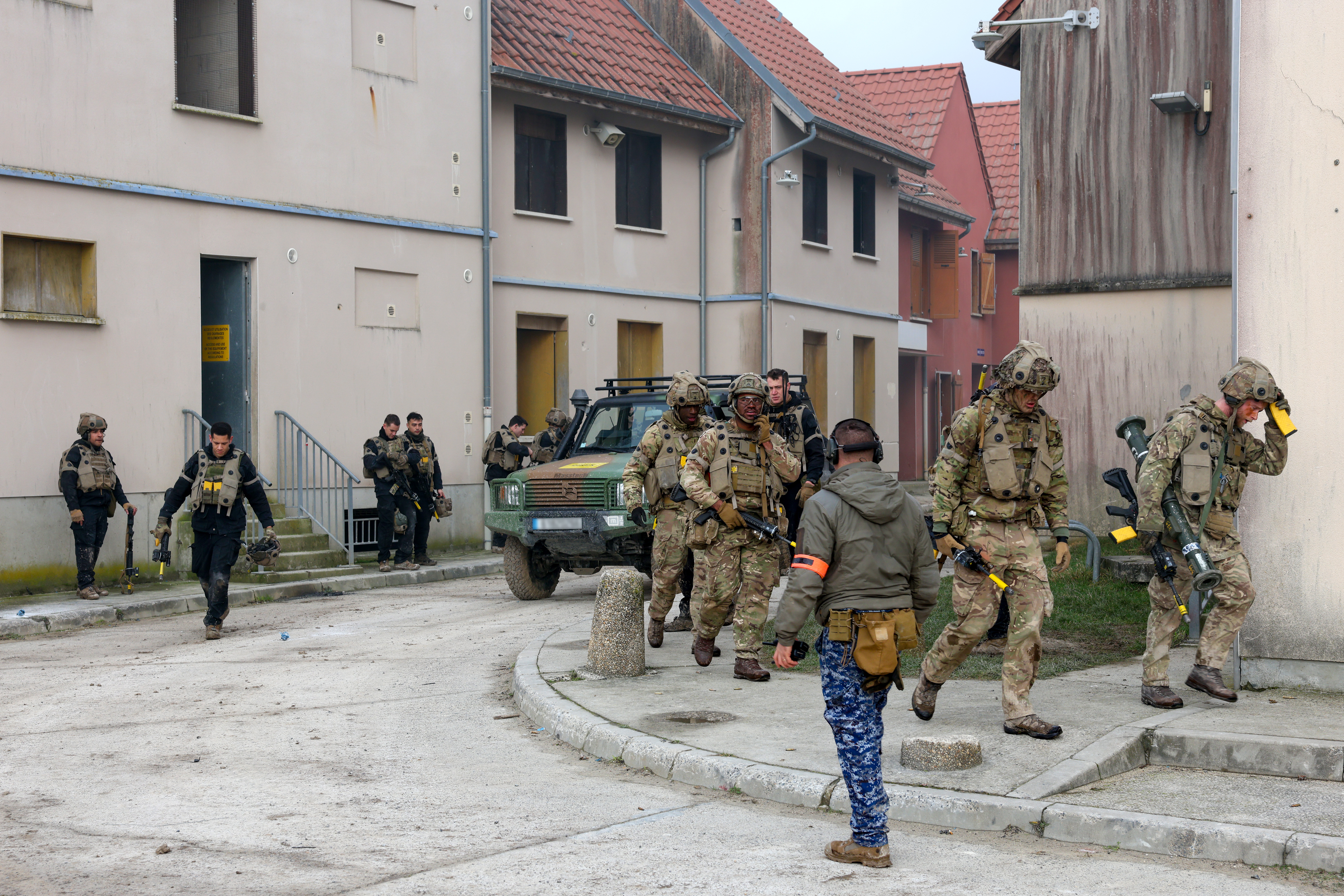 Group of soldiers in tactical gear moving through a residential training area with a military vehicle nearby.