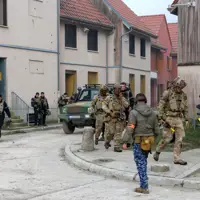 Group of soldiers in tactical gear moving through a residential training area with a military vehicle nearby.