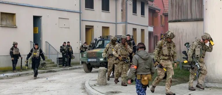 Group of soldiers in tactical gear moving through a residential training area with a military vehicle nearby.