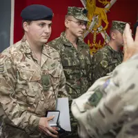 Soldier in camouflage uniform holding an award box during a military ceremony with fellow soldiers standing in line.
