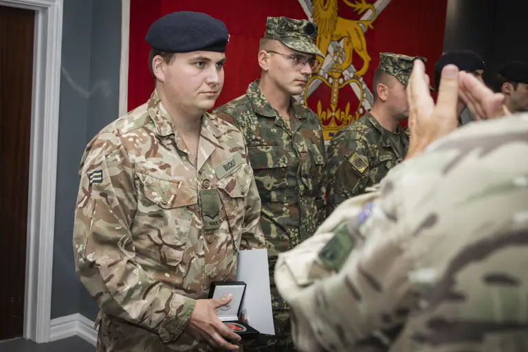 Soldier in camouflage uniform holding an award box during a military ceremony with fellow soldiers standing in line.