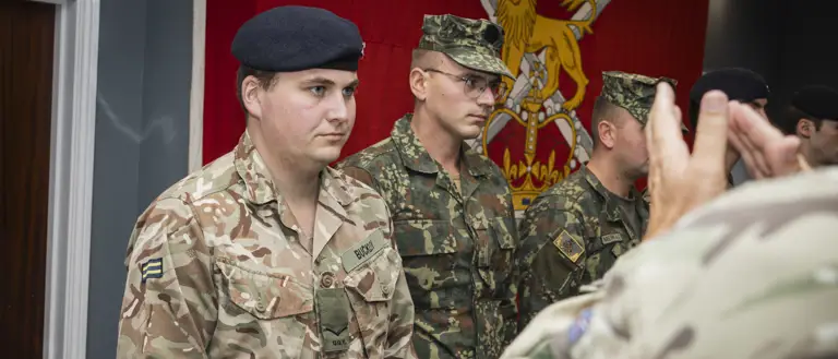 Soldier in camouflage uniform holding an award box during a military ceremony with fellow soldiers standing in line.