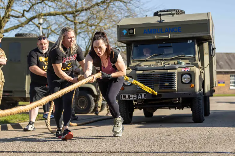 A woman intensely pulls a rope attached to a military ambulance, assisted by a guide. Spectators watch as she demonstrates strength and determination.