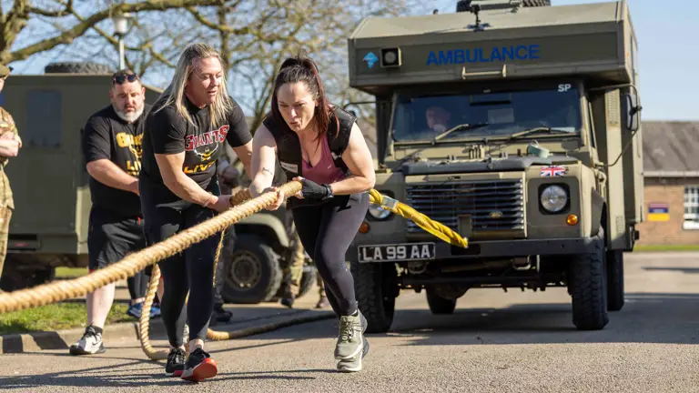 A woman intensely pulls a rope attached to a military ambulance, assisted by a guide. Spectators watch as she demonstrates strength and determination.